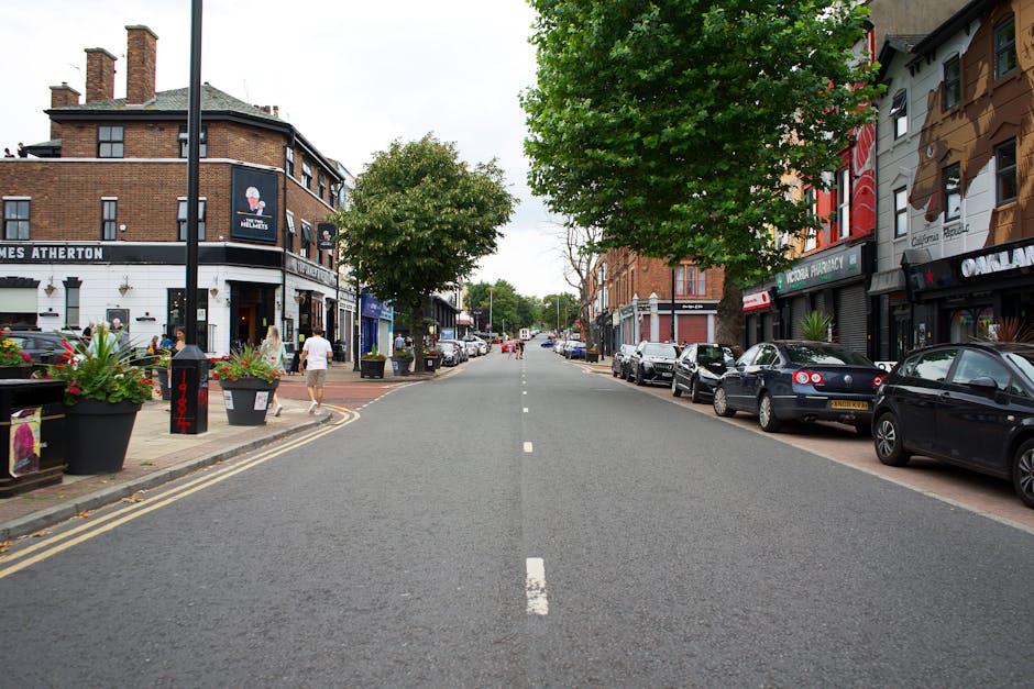 A straight view of a residential street in Anerley Hill, Crystal Palace, showing parked cars lining both sides of the road and storefronts with signage and large flower pots on the pavement. The street features a central white dashed line, with some pedestrians walking along the sidewalk. On the left, there is a two-story building with a white ground floor housing a pub or café, decorated with hanging plants, while on the right, a row of shops with signs such as a pharmacy and other retail outlets is visible. A mid-sized green tree extends overhead, providing some shade, and the scene is captured during daytime with natural lighting. This location is relevant to house removals and furniture transport, with potential considerations for parking and access during a home relocation, which Crystal Palace Removals specializes in, particularly for narrow access streets like Anerley Hill.