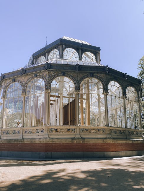 A large Victorian-style glass greenhouse structure featuring multiple domed roofs with intricate metal framework, situated outdoors against a clear sky. The greenhouse has prominent arched windows and