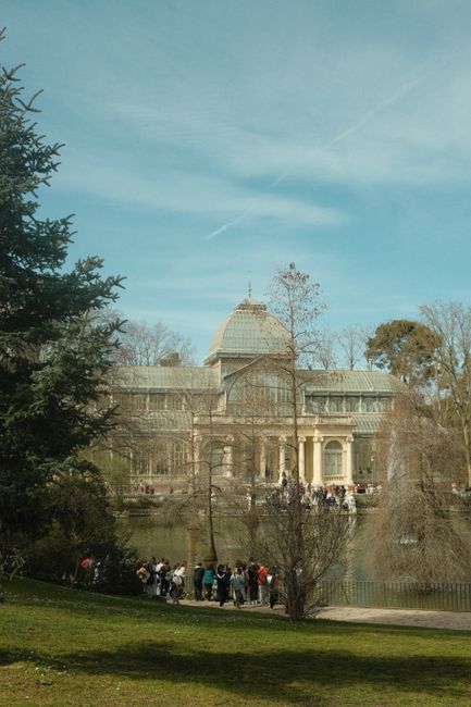 The image shows a large historic glass and stone building with a domed roof, situated in a park-like setting with trees and a pond in the foreground. A group of people are gathered near the pond, poss