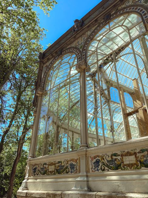 The image shows the exterior of an ornate glasshouse with a decorative painted base, situated outdoors amidst tall green trees under a bright blue sky. The glass structure features large, clear glass panels framed by white metal supports, allowing sunlight to illuminate the interior and create reflections on the glass. The ornate painted base of the glasshouse includes intricate patterns and motifs, adding to its historic and decorative appearance. The setting appears to be part of a garden or park environment, with nearby trees casting dappled shadows on the structure. This type of glasshouse is commonly used for botanical displays or entertainment venues, and the image likely relates to house removals or moving services focused on relocating large, fragile glass or decorative architectural elements, as offered by Crystal Palace Removals.