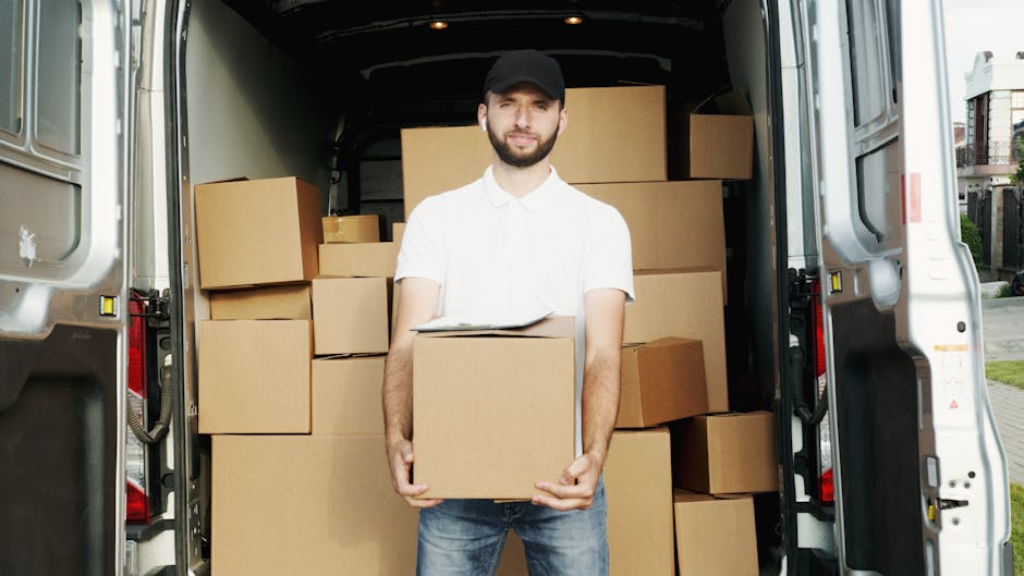 A man with a beard, wearing a white short-sleeved shirt, blue jeans, and a black cap, stands inside the open rear of a moving van, holding a cardboard box in front of him during a house relocation process. Behind him, the van's interior is filled with multiple cardboard boxes of varying sizes, stacked neatly in the cargo area, some wrapped in plastic or secured with packing materials. The van is parked on a residential street near a sidewalk, with a house and greenery visible in the background. The image captures the loading process of furniture transport and packing, associated with professional removals services by Crystal Palace Removals, illustrating the careful handling and organization involved in home moves.
