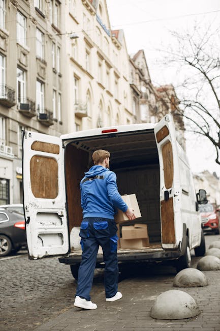 A man with light brown hair, dressed in a blue jacket, matching blue trousers, and white sneakers, is standing at the open rear doors of a white cargo van parked on a cobblestone street in an urban area. Inside the van, several cardboard boxes wrapped in plastic and brown paper are visible, with some being carried by the man towards the building. The van is positioned near a curb with smooth, rounded stone bollards separating it from the sidewalk. The street is lined with multi-storey Victorian-style buildings with cream facades, large windows, and small balconies, suggesting a residential area in Crystal Palace. Bare tree branches are visible against a cloudy sky, indicating a colder season. Nearby cars are parked along the street, and the scene appears to be part of a home relocation or furniture transport process managed by Crystal Palace Removals, supporting professional packing and moving services.
